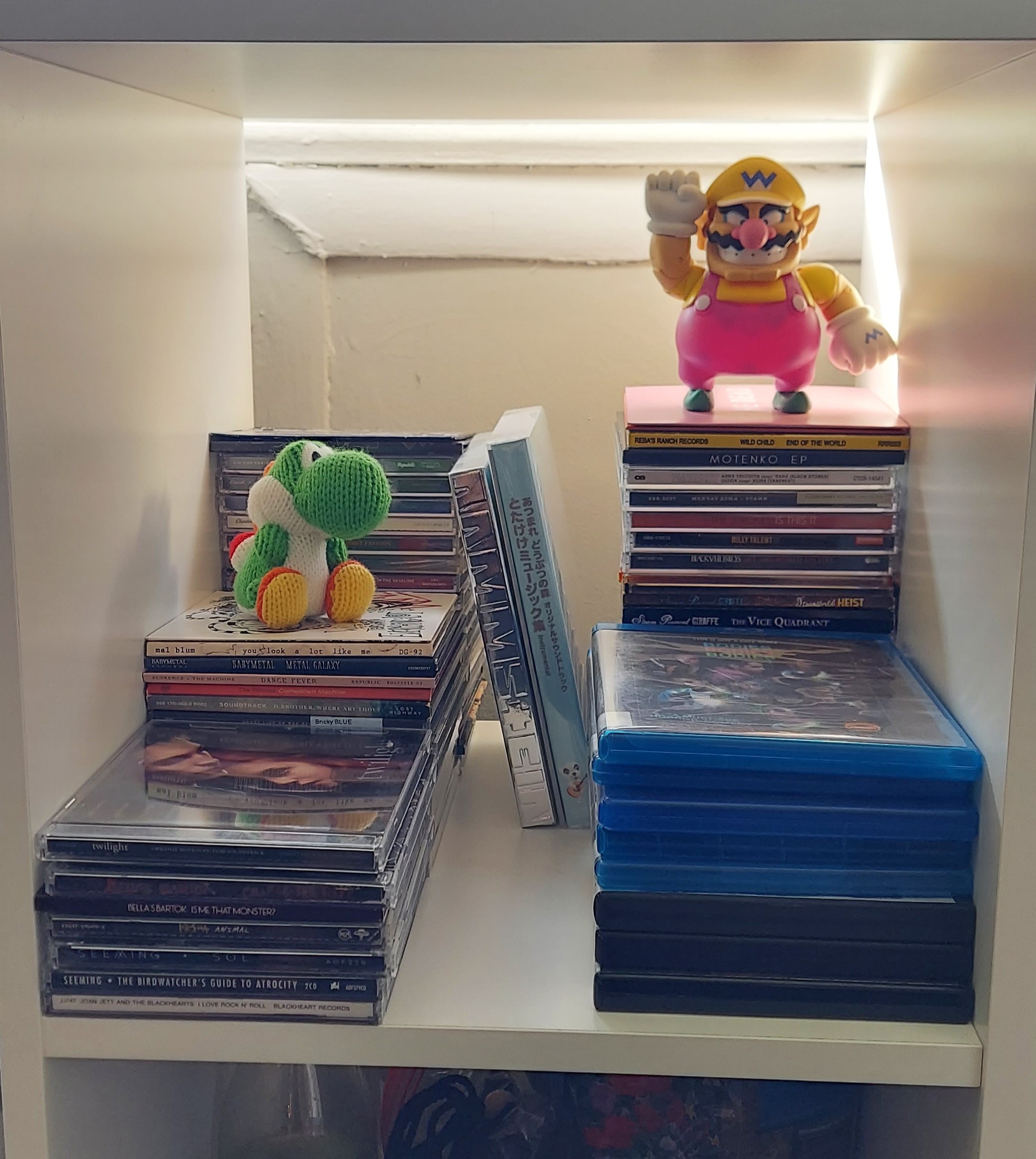 white square shelf with stacks of CDs arranged on it. A stack of Blu-rays is set off to the right side of the shelf. A Yarn Yoshi amiibo and a Wario figure sit atop two of the CD stacks.